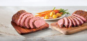 Cooked Corned Beef sliced on a tray with a plate of carrots and potatoes in the background.